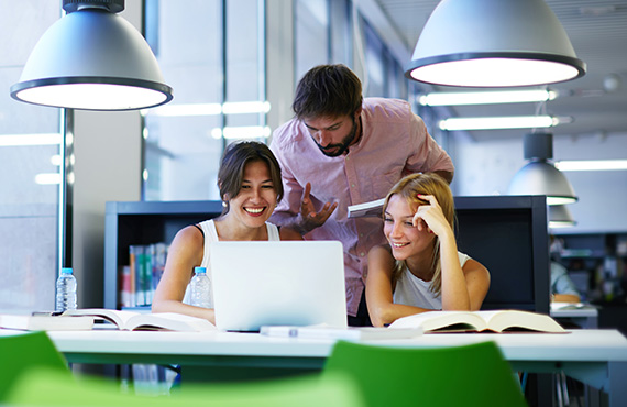 Group of international university students having fun studying in library, three colleagues of modern work co-working space talking and smiling while sitting at the desk table with laptop computer